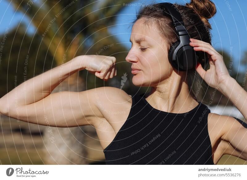 Young beautiful woman in sportswear raises arms to show muscles, biceps, demonstrates strong hand, has slim figure outdoors, feels confident in victory, looks independent, smiles positively