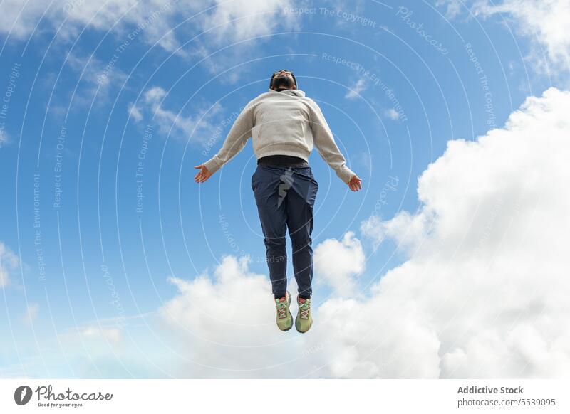 Unrecognizable man jumping against cloudy blue sky in daylight enjoy happy hands apart carefree summer nature male sneakers freedom travel energy casual excited