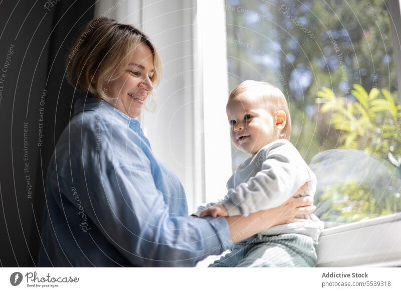 Mature blond woman and a baby boy near the window at home mother infant child love care childcare parent motherhood childhood family room parenthood bonding