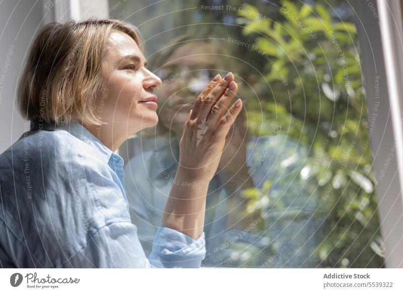 Mature blond woman near the window at home care room apartment light tranquil daylight living room indoors blonde mature female tree reflection side view alone