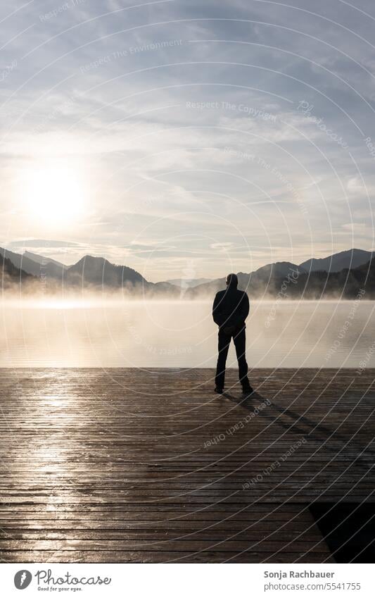 A man alone on a wooden jetty by the lake. Sunrise, autumn, back view. Man Rear view Silhouette Lake wooden walkway Stand Morning Fog Autumn Water Calm