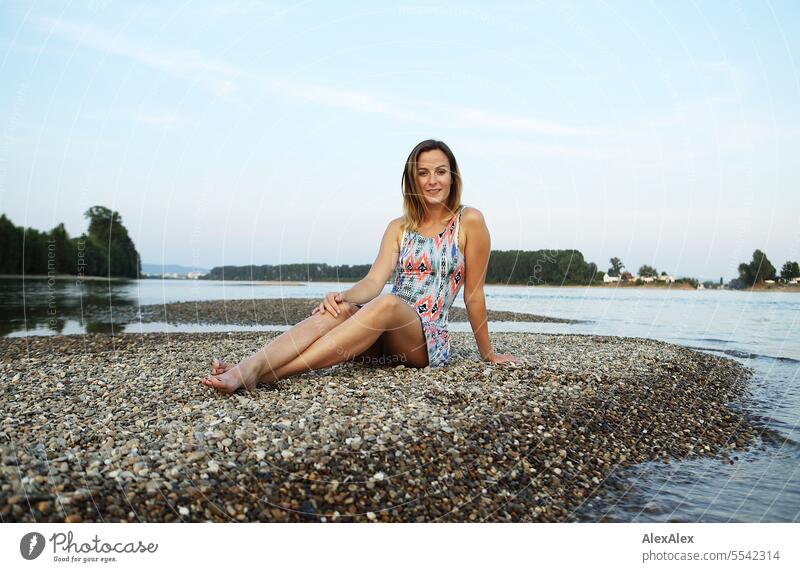 A beautiful, slim, young woman in a summer dress sits barefoot on a sandbank of gravel in the Rhine and looks into the sunset Woman beautiful woman Slim