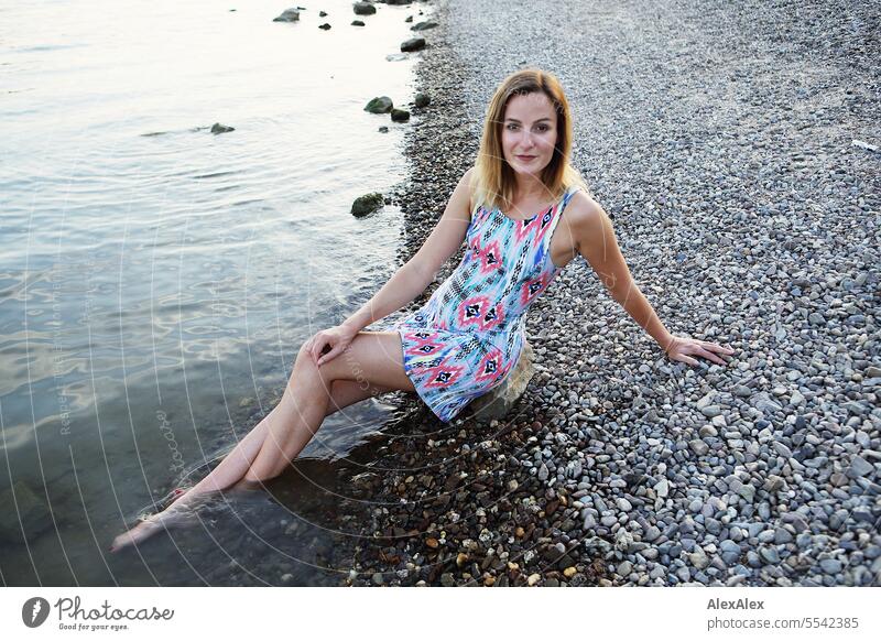 Beautiful slim young woman in summer dress sits barefoot on Rhine shore made of gravel with feet in water Woman beautiful woman Slim Athletic youthful teen