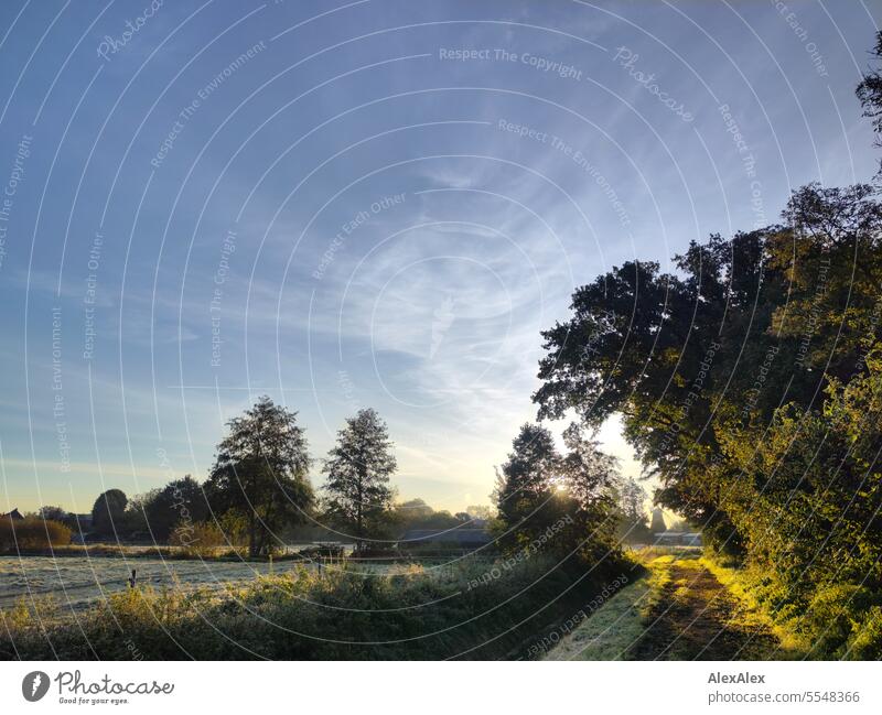 Sunrise/ morning atmosphere in early autumn, a dirt road to fenced pastures with drainage ditch trees, houses and a windmill Morning in the morning Moody