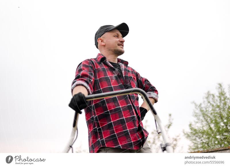 Bottom view of tired man makes a short break while cutting grass with a lawn mower after at backyard of his house. Gardening, sweating or exhausted male resting after working outdoor household chores