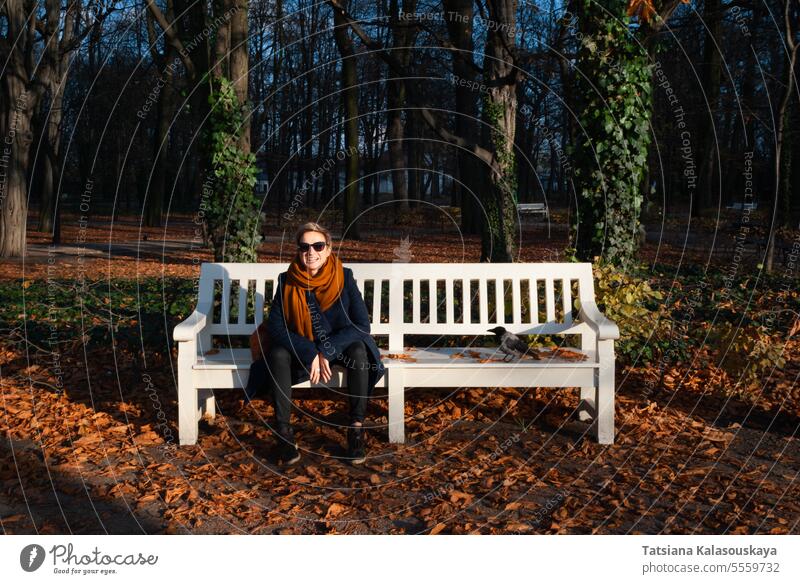 A short-haired woman sits on a white bench near a crow in autumn park female fall Hooded crow Corvus cornix scald-crow bird people person adult Blond short hair