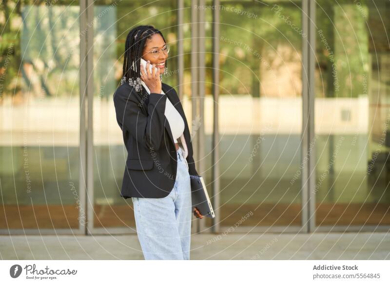 Smiling businesswoman walking and talking on mobile phone while holding laptop smartphone entrance speak smile entrepreneur modern office doorway using