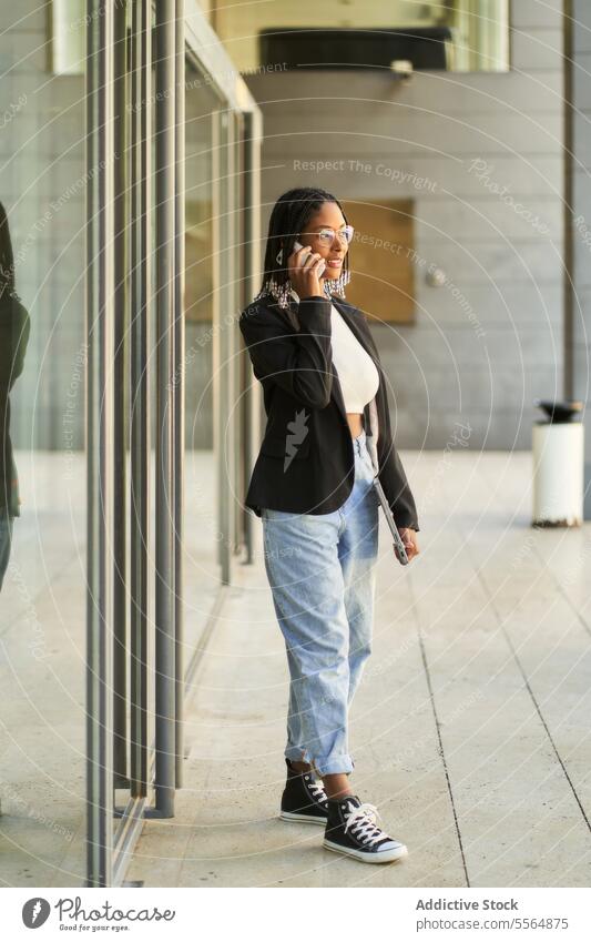 Smiling businesswoman walking and talking on mobile phone while holding laptop smartphone entrance speak smile entrepreneur modern office doorway using