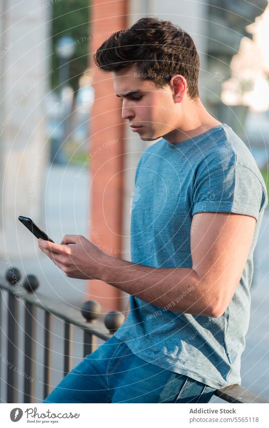 Young man with blue t-shirt using smartphone in city. caucasian young Gen-Z railing leaning engrossed street hair brunette casual jeans denim afternoon vertical