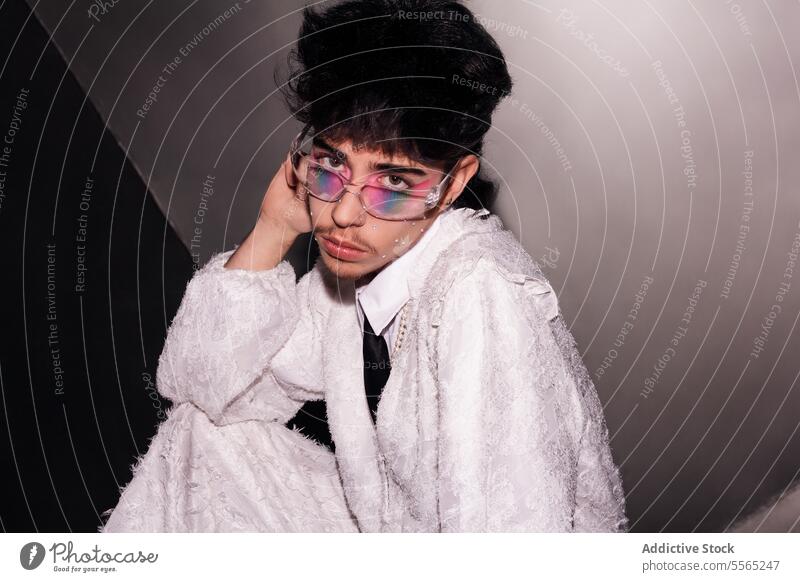 Man Daring gaze in a radiant suit. individual makeup sitting concrete floor white camera dual-tone wall fashion-forward strong trend edgy style glasses tie lace