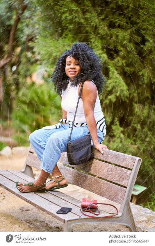 woman in Relaxed moment in nature green black outdoors summer african curly hair park bench casual bag smartphone day serene trees greenery sitting relaxation