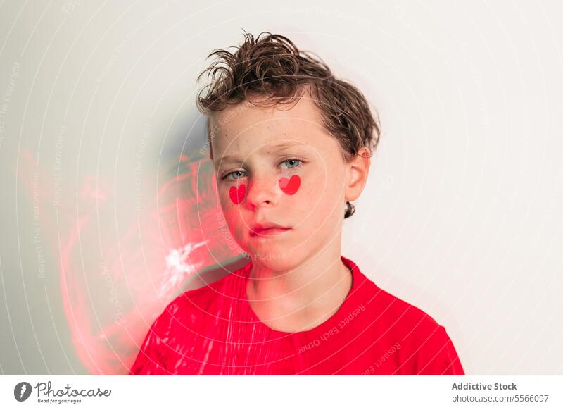 Calm boy with heart stickers on cheeks and abstract red patterns Boy curly hair calm gaze red shirt white background emotion child young expression love serene