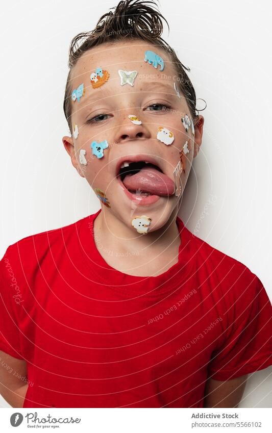 Boy with colorful stickers on his face and wet hair tongue animal playful child young expression red shirt white background fun silly emotion cheerful cute kid