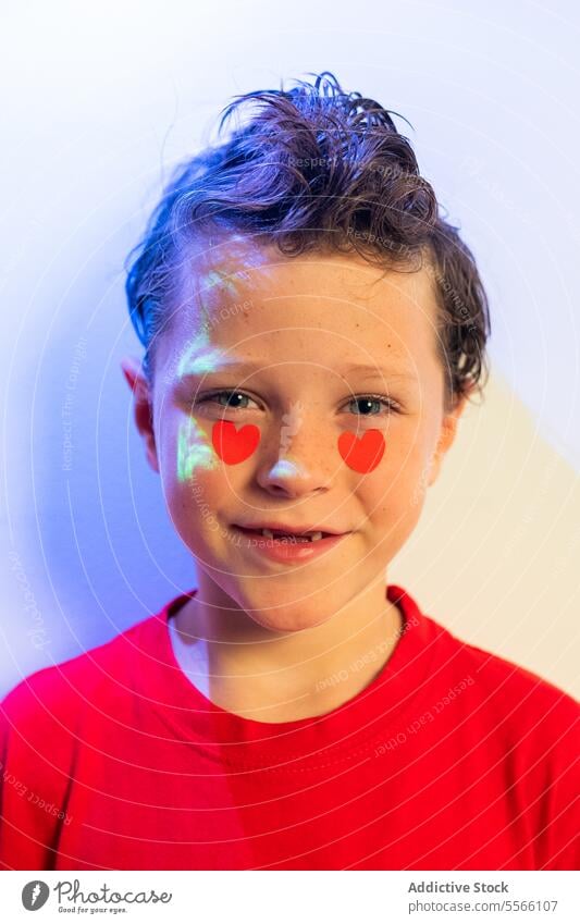 Calm boy with heart stickers on cheeks and abstract red patterns Boy curly hair calm gaze red shirt white background emotion child young expression love serene