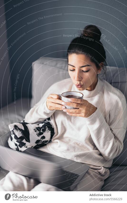 Crop young woman sitting with laptop and coffee cup using internet connection online living room at home female dark hair sofa gadget device beverage netbook