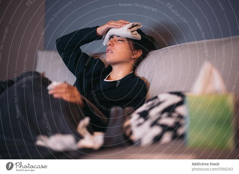 Young woman under weather resting on sofa in living room towel lying relax interior sick napkin eyes closed at home female young blanket warm clothes dark hair