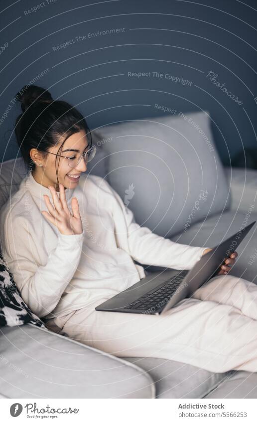 Happy young woman resting on sofa and attending video call on laptop at home hello gesture smile wave hand living room female greeting eyeglasses toothy smile