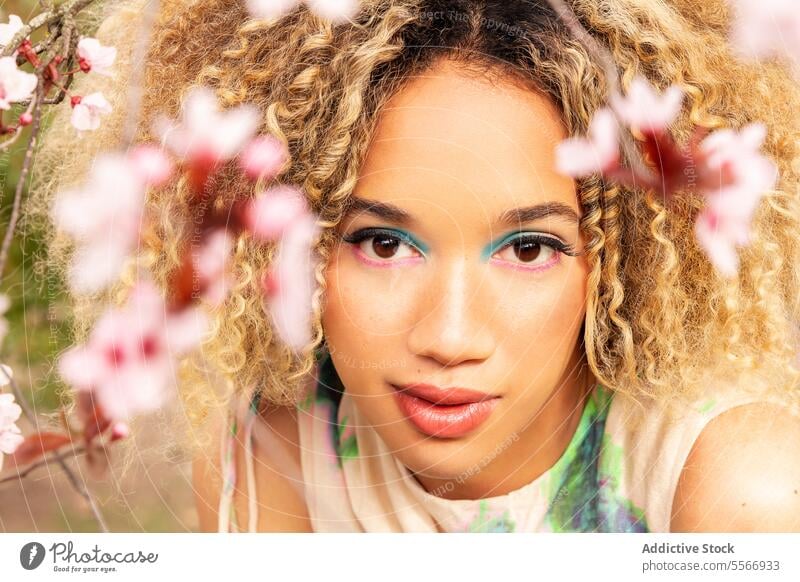 Ethnic woman amidst pink flowers in patterned dress Woman blonde curly hair playful mysterious nature camera gaze field outdoors environment portrait look stand