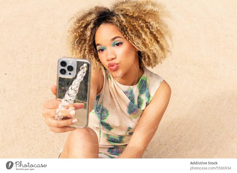 Ethnic woman taking selfie against beige background Woman phone curly hair blonde turquoise eyeshadow pink lips pucker modern smartphone glittery case camera