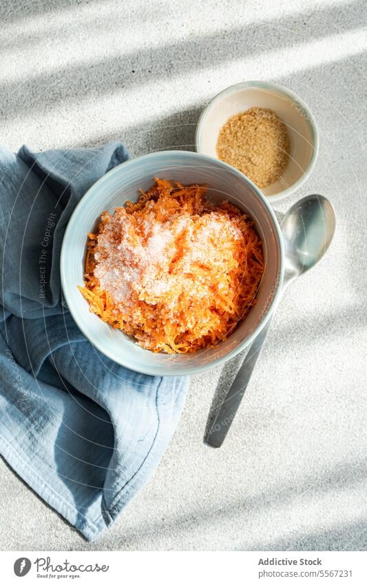 Carrot dessert with chopped raw carrot vegetable with sugar served on bowl on table blue grated spoon cloth gray beige sunlight shadow texture close-up fresh