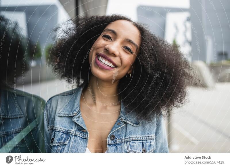 Ethnic young woman smiling and leaning against a reflective glass pane curly hair denim jacket white top smile reflection joy street urban fashion casual style