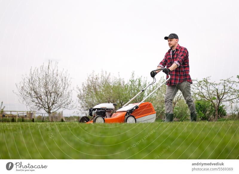 view of man in casual clothes mows lawn with lawn mower at backyard of his house. Husband takes care of garden on spring cloudy day. Modern gasoline garden equipment. Landscaping work