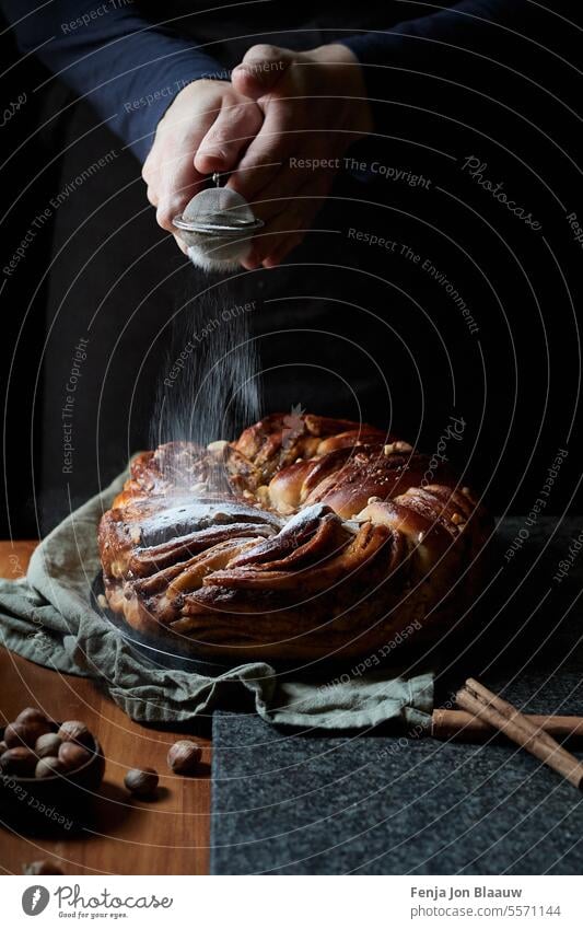 Freshly baked cinnamon loaf, with human hands sprinkling icing sugar on top in a moody setting in the kitchen action baking cinnamon bread dark and moody