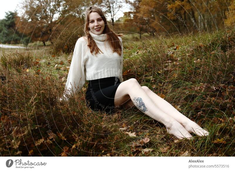 Young woman in white wool sweater and black skirt sits barefoot in a northern German heath landscape in the fall in the grass between heather and smiles Woman