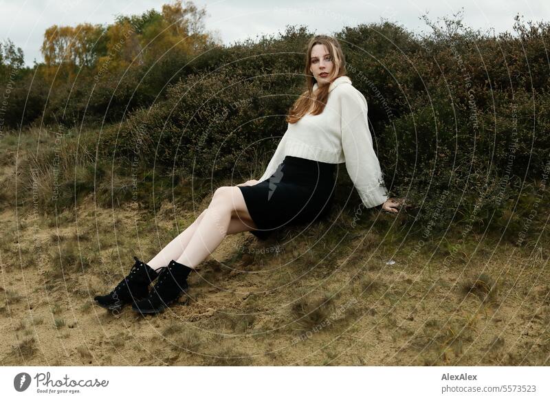 Young woman in white wool sweater and black skirt sits in the grass between heather in a northern German heath landscape in fall Woman Large tall woman