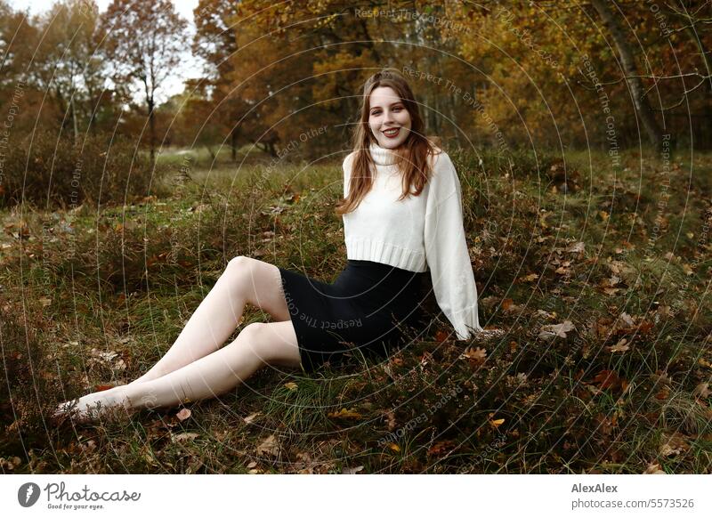 Young woman in white wool sweater and black skirt sits barefoot in a northern German heath landscape in the fall in the grass between heather and smiles Woman