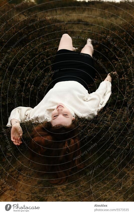 Young woman in white woolen sweater and black skirt lying upside down on a rampart in the heath between grass and heather Woman Large tall woman long legs Slim