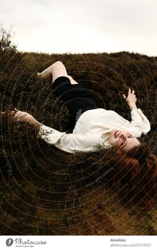 Young woman in white woolen sweater and black skirt lying upside down on a rampart in the heath between grass and heather Woman Large tall woman long legs Slim