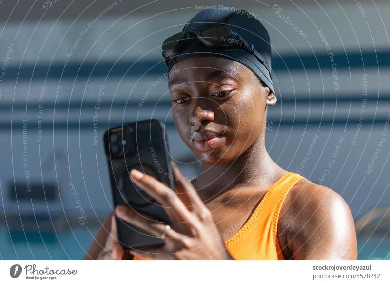 Black young woman holding her smartphone while standing at the swimming pool black female heated swimsuit orange healthy smiling blue lifestyle real people