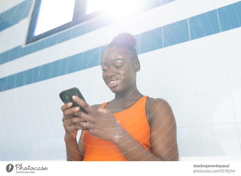 Black young woman using her smartphone under a beautiful light at the swimming pool black female heated swimsuit orange healthy blue lifestyle sport real people