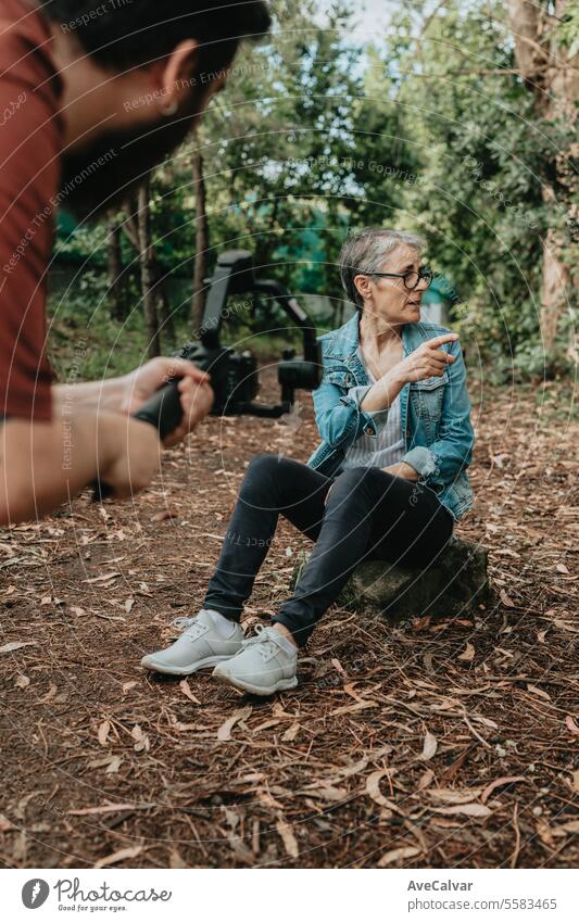 Senior jubilant woman explaining her trip to followers while a guy records her live on social media. senior retirement streamer modern female person nature