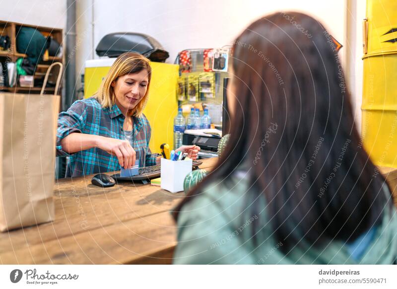 Female cashier passing credit card through store terminal to charge purchased items by customer female shop pc computer local smile happy assistant finish