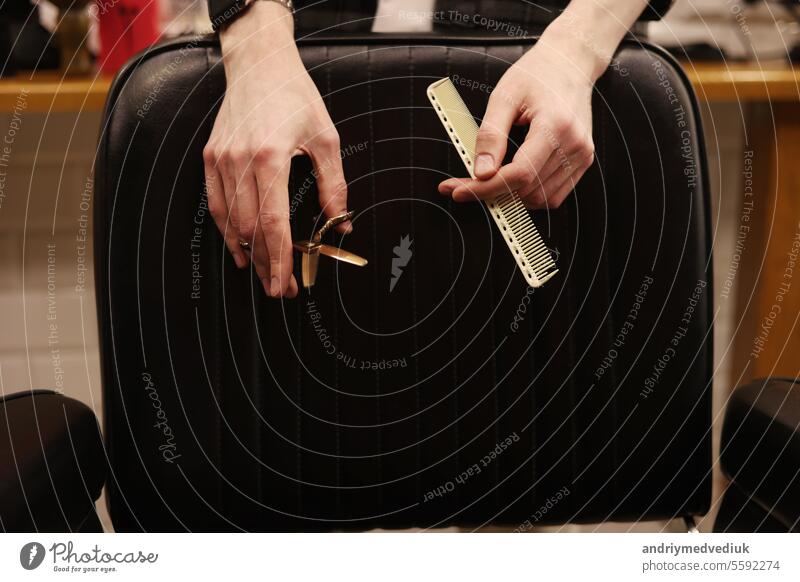 Hair style, hairdresser, beauty and customer service concept. Cropped shot of barber man hands hold professional stylish gold hairbrush and scissors by the workplace, leather black chair in barbershop