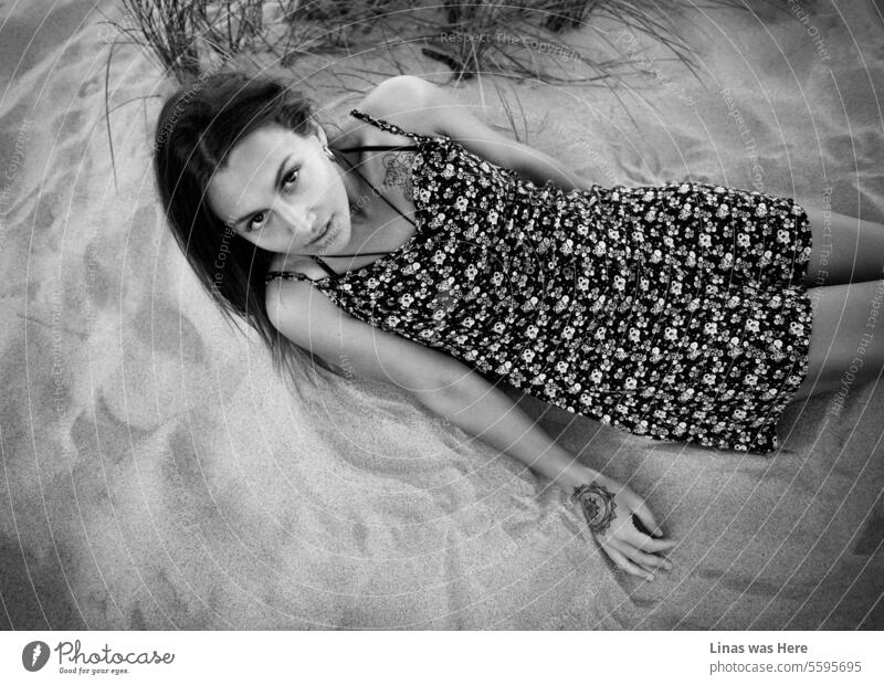 A stunning brunette is reclining on the sandy expanse of these untamed dunes, flawlessly showcasing her modeling prowess. An enchanting woman graces the scene in a breezy summer dress, gazing directly into the camera.