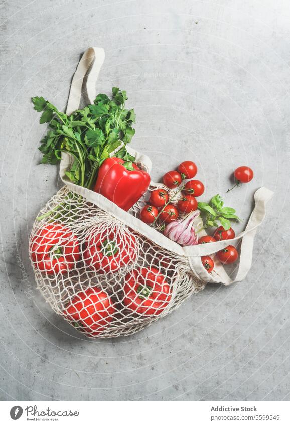 Top view of various vegetables and herbs in reusable plastic free shopping bag at grey concrete kitchen table red bell pepper tomatoes cilantro garlic