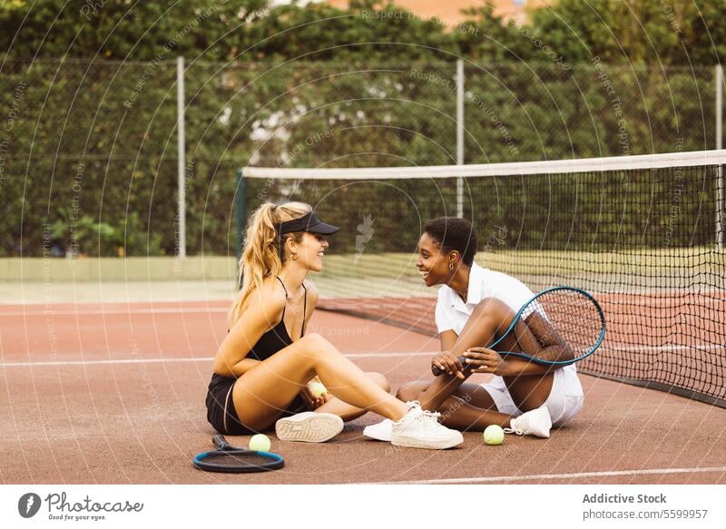 Two young beautiful women having a conversation sitting on a tennis court next to the net. Two amateurs tennis players having a break during a tennis match.