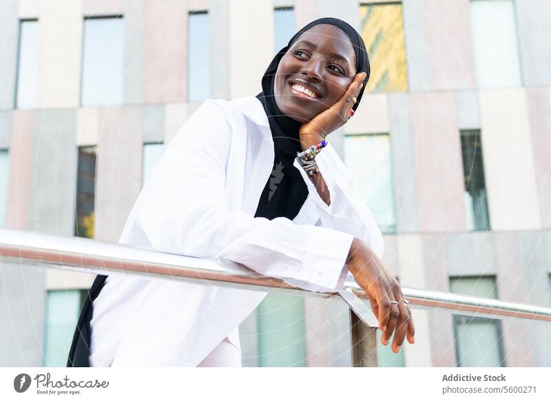 From below cheerful African American Muslim businesswoman in black hijab touching chin and looking away while leaning on railing in city african american muslim