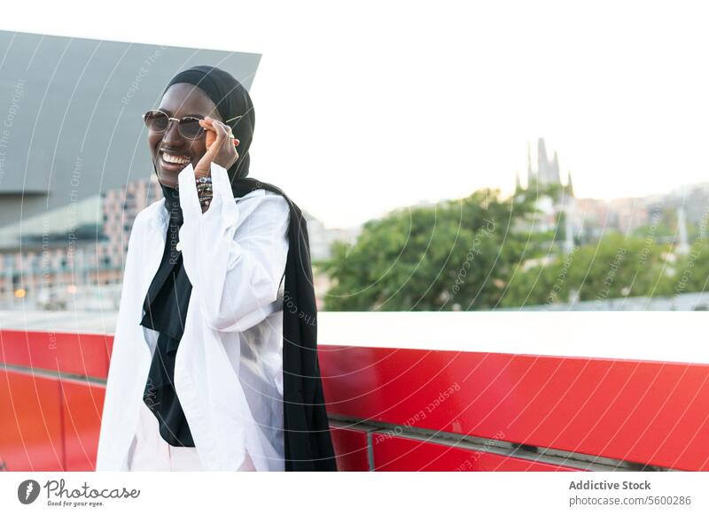 Happy entrepreneur in hijab wearing goggles in city businesswoman sunglasses stylish cheerful muslim tradition black headscarf standing retaining wall sky smile