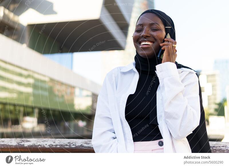 Happy woman in hijab talking over mobile phone in city businesswoman smartphone eyes closed cheerful modern building standing african american cellphone formal