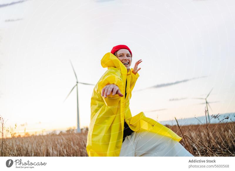 Smiling woman in yellow dancing near wind turbines raincoat red beanie field sunset dance joy vibrant nature renewable energy eco-friendly enjoyment happy fun