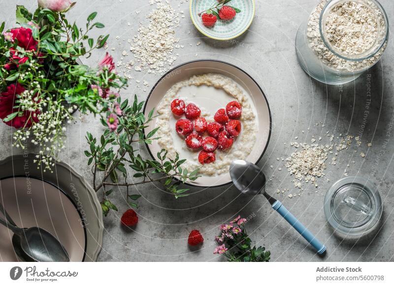 Bowl with porridge and heart shaped raspberries at grey concrete kitchen table with glass jar, branches, bowls and spoon. Preparing healthy vegan breakfast at home. Top view.