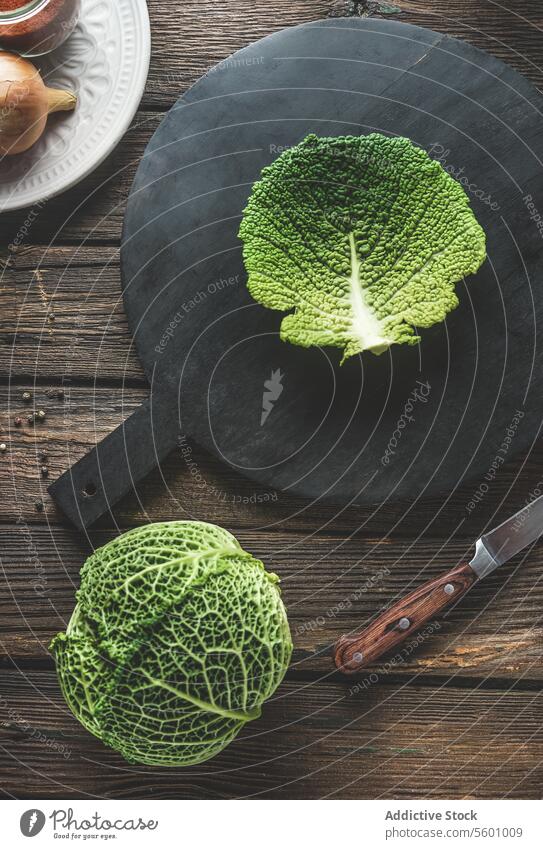 Whole raw savoy cabbage and cabbage leaf on black cutting board on rustic wooden kitchen table with knife. Preparing food with seasonal autumn vegetable. Top view