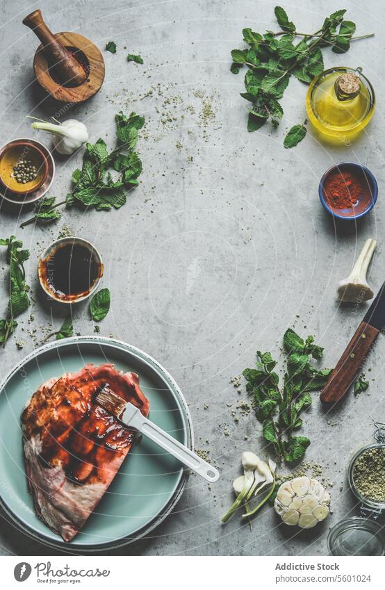 Raw marinated lamb on plate at kitchen table with mint, spices, garlic, oil, mortar and pestle and kitchen utensils. Preparing bbq meat at home with red marinade. Top view . Frame