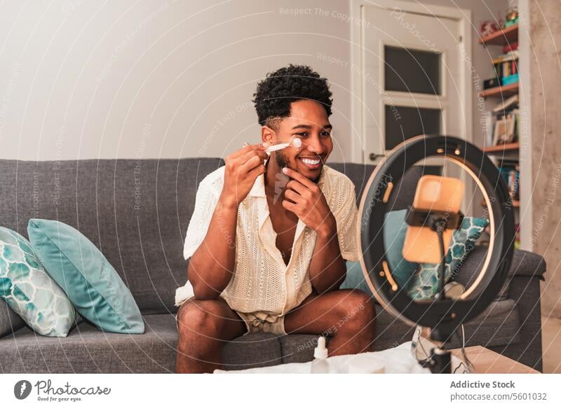 A cheerful young latin man applies facial roller in a well-lit living room, reflecting a modern self-care lifestyle for video blog streaming skincare cream