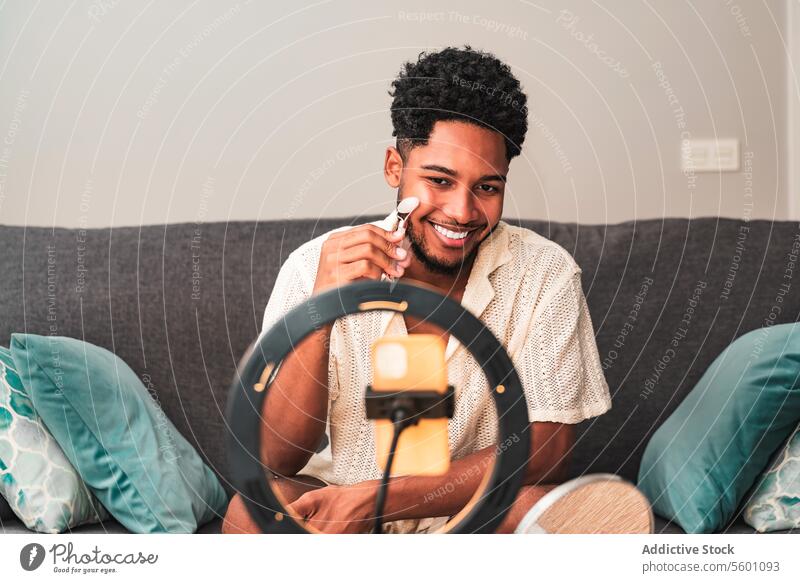 A cheerful young latin man records a skin care theme video blog on his smartphone secured to a ring light, smiling while using a facial roller on a couch at home.