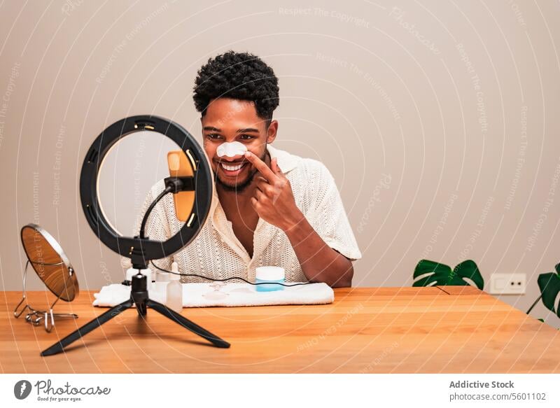 A cheerful latin man is applying cream to his face while sitting at a table with a skincare routine set-up, including a mirror with an integrated ring light and various skincare products.